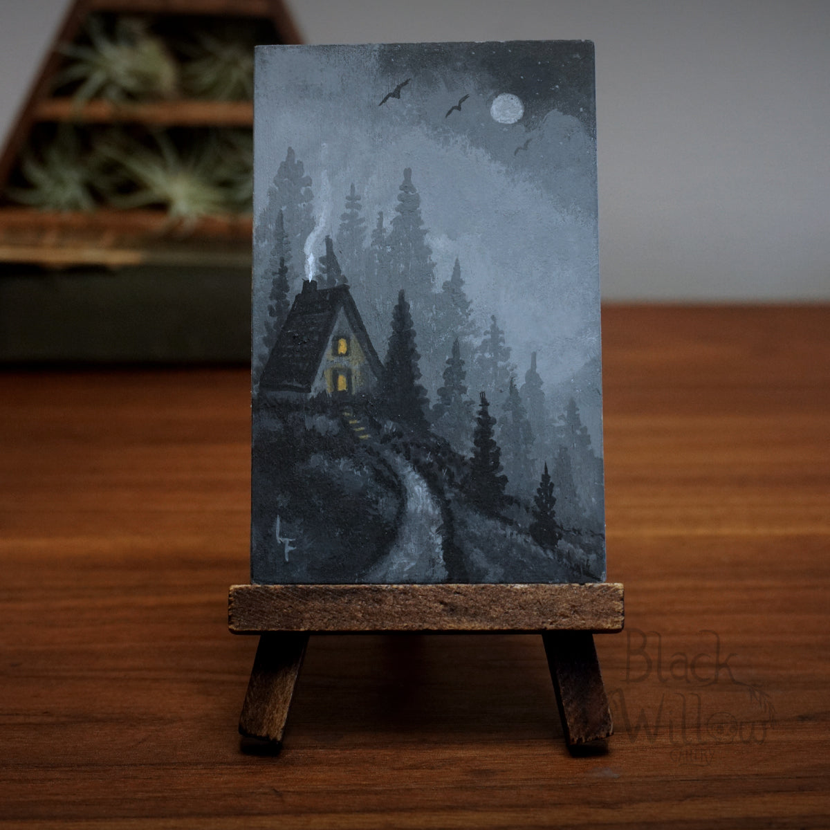 Miniature painting of a cabin and crows passing in the distance in the woods on a small easel