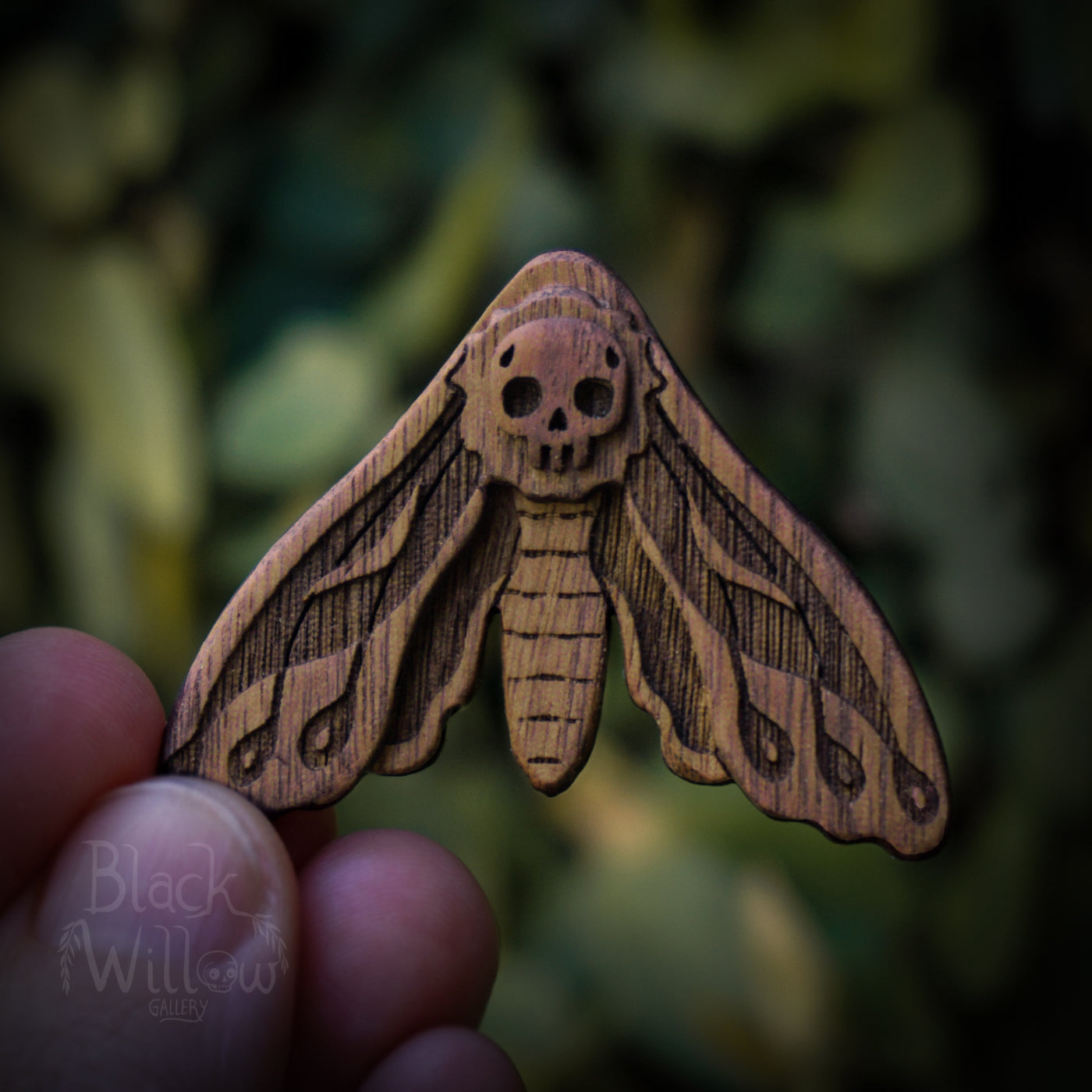 Wooden moth-shaped pin with skull design held by a hand against a blurred natural background.
