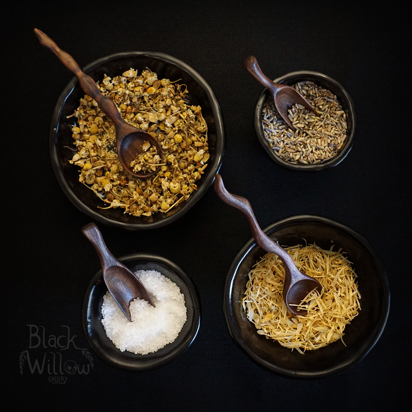 Four small dark bowls containing loose herbal tea ingredients,aech one with a wooden spoon  including dried flowers, shredded yellow herbs, white salt, and lavender, arranged on a black background with wooden spoons. A 'Black Willow Gallery' logo is visible in the bottom left corner.