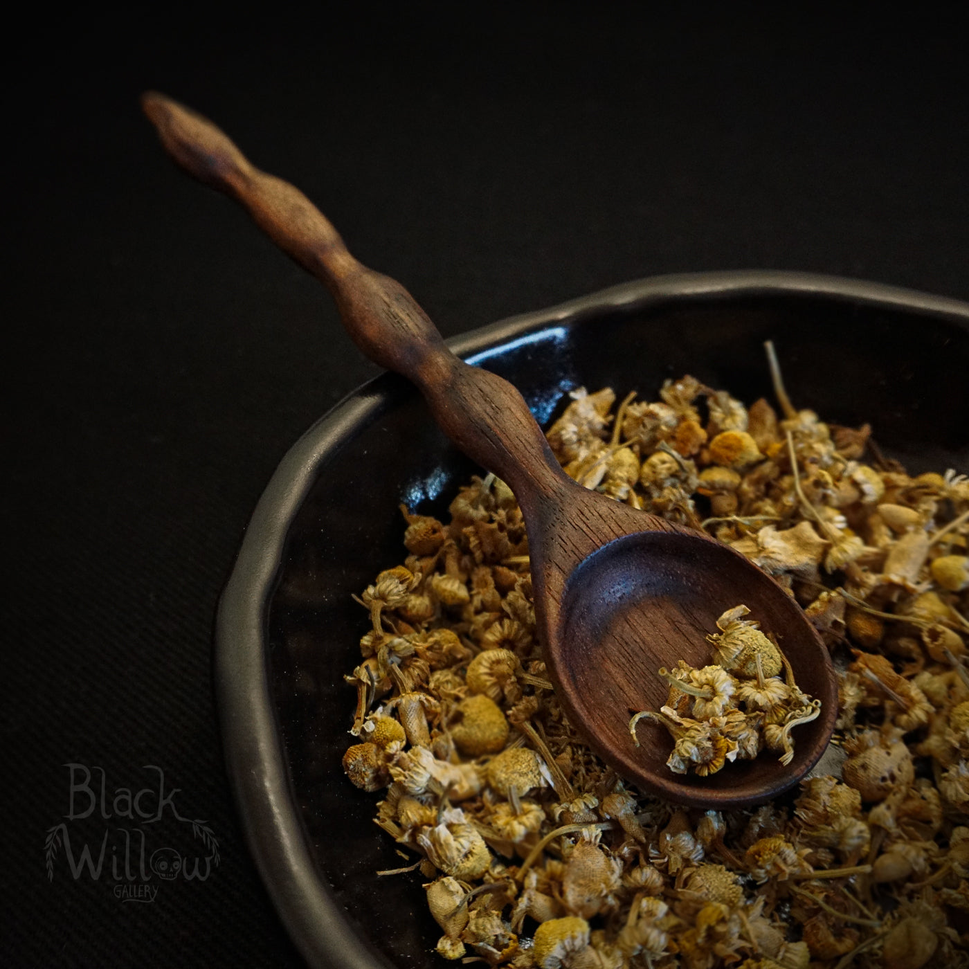 A close-up of dried chamomile tea in a dark bowl with a wooden spoon, set against a dark background. The brand name 'Black Willow Gallery' is visible as a watermark in the bottom-left corner.