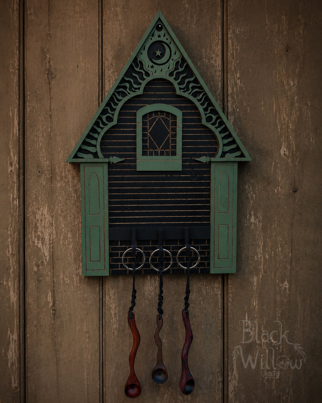 A decorative black cuckoo clock shaped like a house with teal shutters and hanging spoon keys, mounted on a rustic brown wooden wall. The brand name 'Black Willow Gallery' is visible in the bottom right corner with 3 twisted wooden spoons keychains