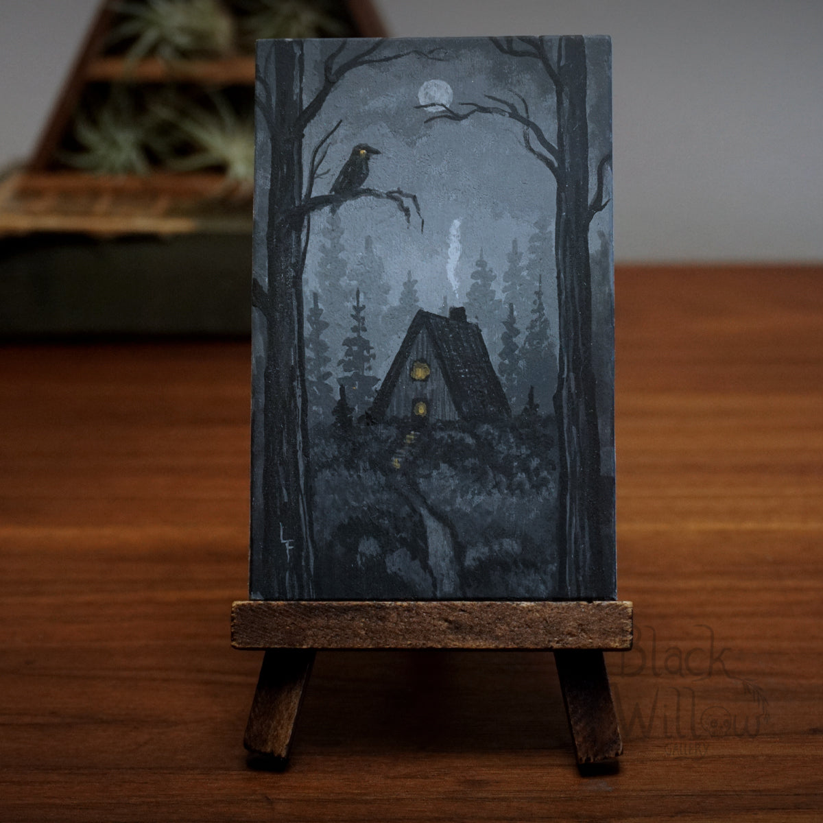 Artistic depiction of a cabin in the woods with a crow on a branch, displayed on a wooden stand.