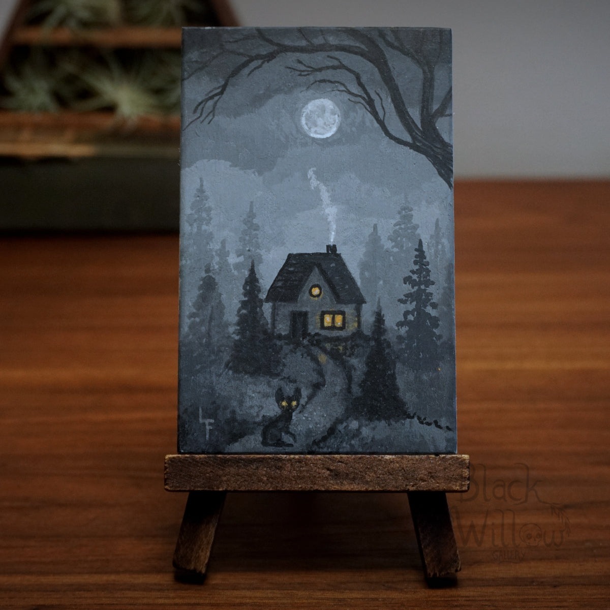 Miniature painting of a house and a cat in a forest with a moonlit sky on a wooden stand.