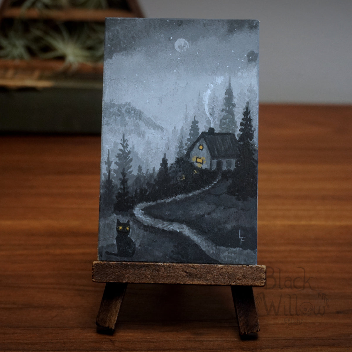 Miniature painting of a house in a forest with a cat on a wooden stand.