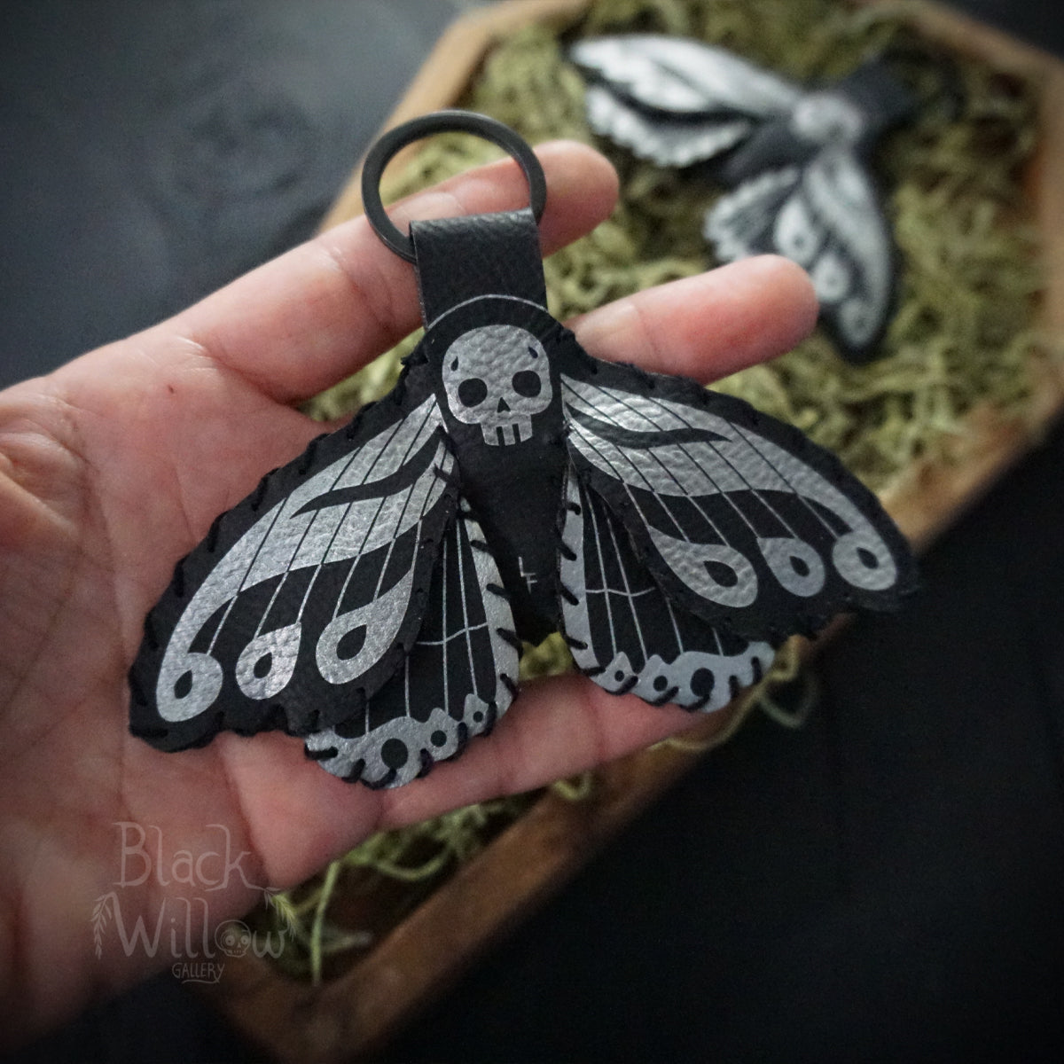 Hand holding a black faux leather death’s-head hawkmoth keychain with silver skull and wing details, with moss and wooden coffin tray in the background.