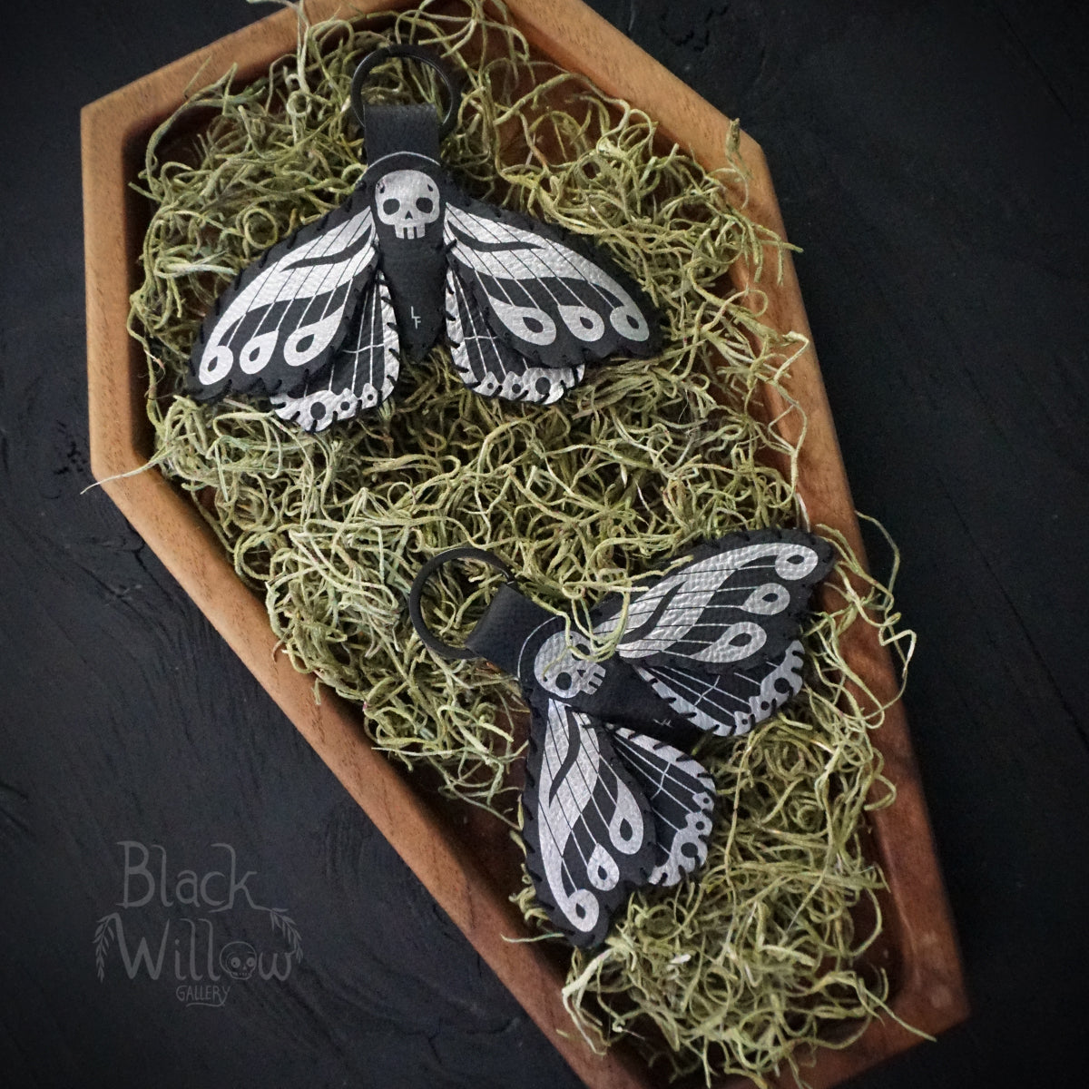 Two black faux leather death’s-head hawkmoth keychains with silver details displayed on green moss inside a wooden coffin-shaped tray.