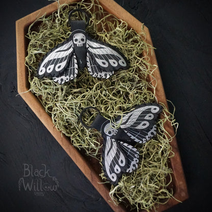 Two black faux leather death’s-head hawkmoth keychains with silver details displayed on green moss inside a wooden coffin-shaped tray.